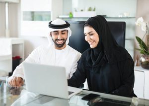 Young Emirati couple is using laptop at home to discuss business matters together. They are using laptop to view statistics and feedback from customers, see results of the development of the company. Both are wearing traditional arabic style clothing. They are in a modern brightly lit living room. Made in Dubai, UAE.